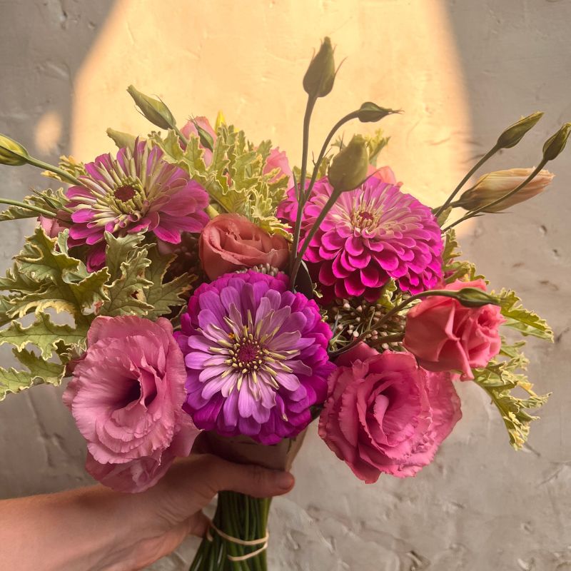 Purple zinnia and pink lisianthus bouquet held in a hand against a sunlit wall
